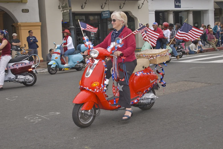 an older woman riding on the back of a red moped with american flags in the back