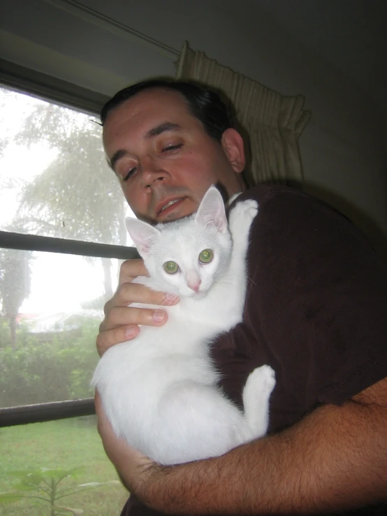 a man holding up his white cat while standing in front of a window