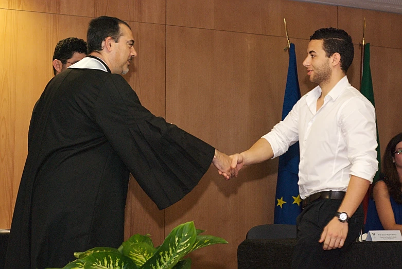 two men in formal clothing shaking hands near a wall