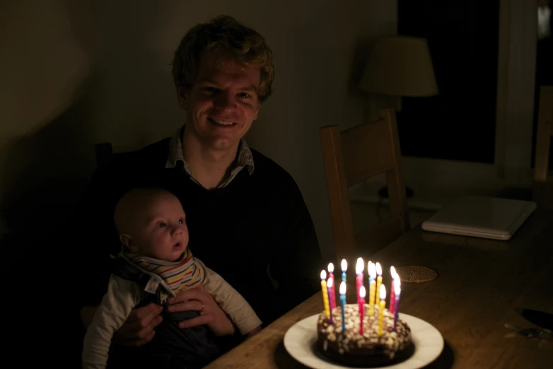 the man is holding a baby near a cake with lit candles