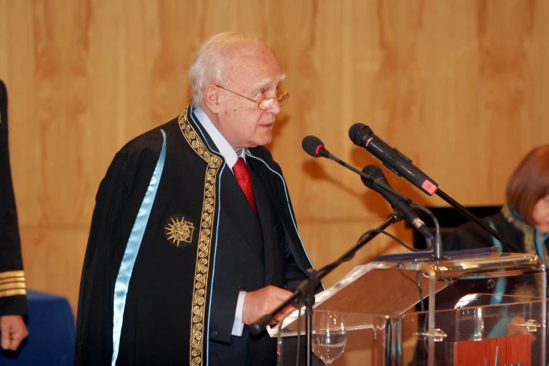 an older man stands at a podium speaking into microphones