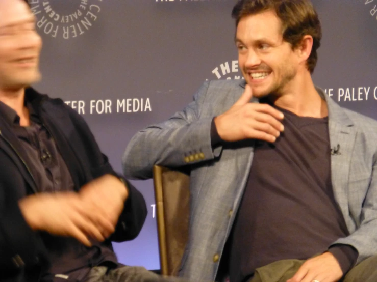 two men smile while sitting in chairs at a panel