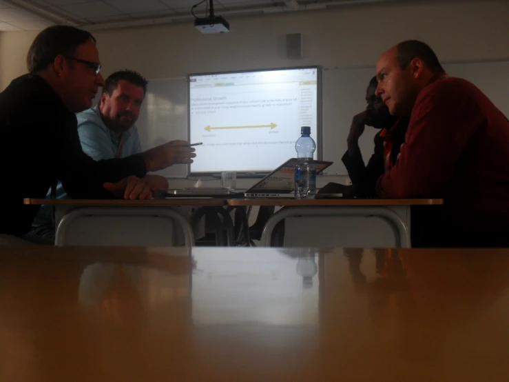 three men at a table looking at a slide presentation on a large screen