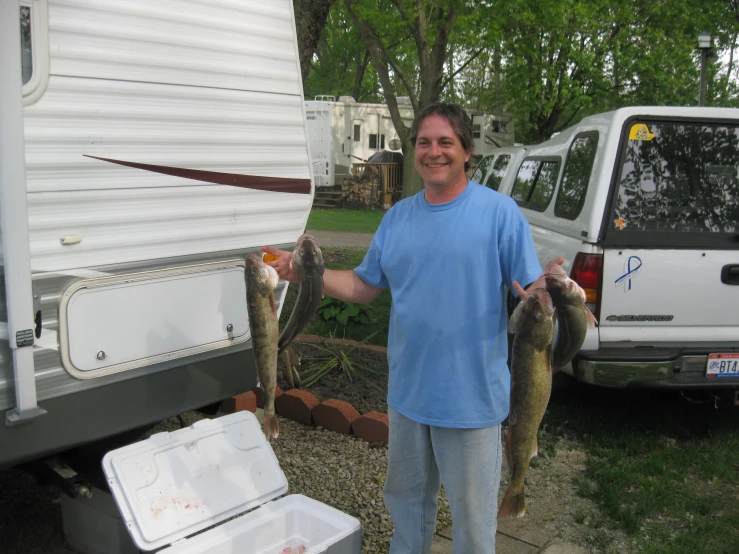 a man holding two fish and standing next to an rv