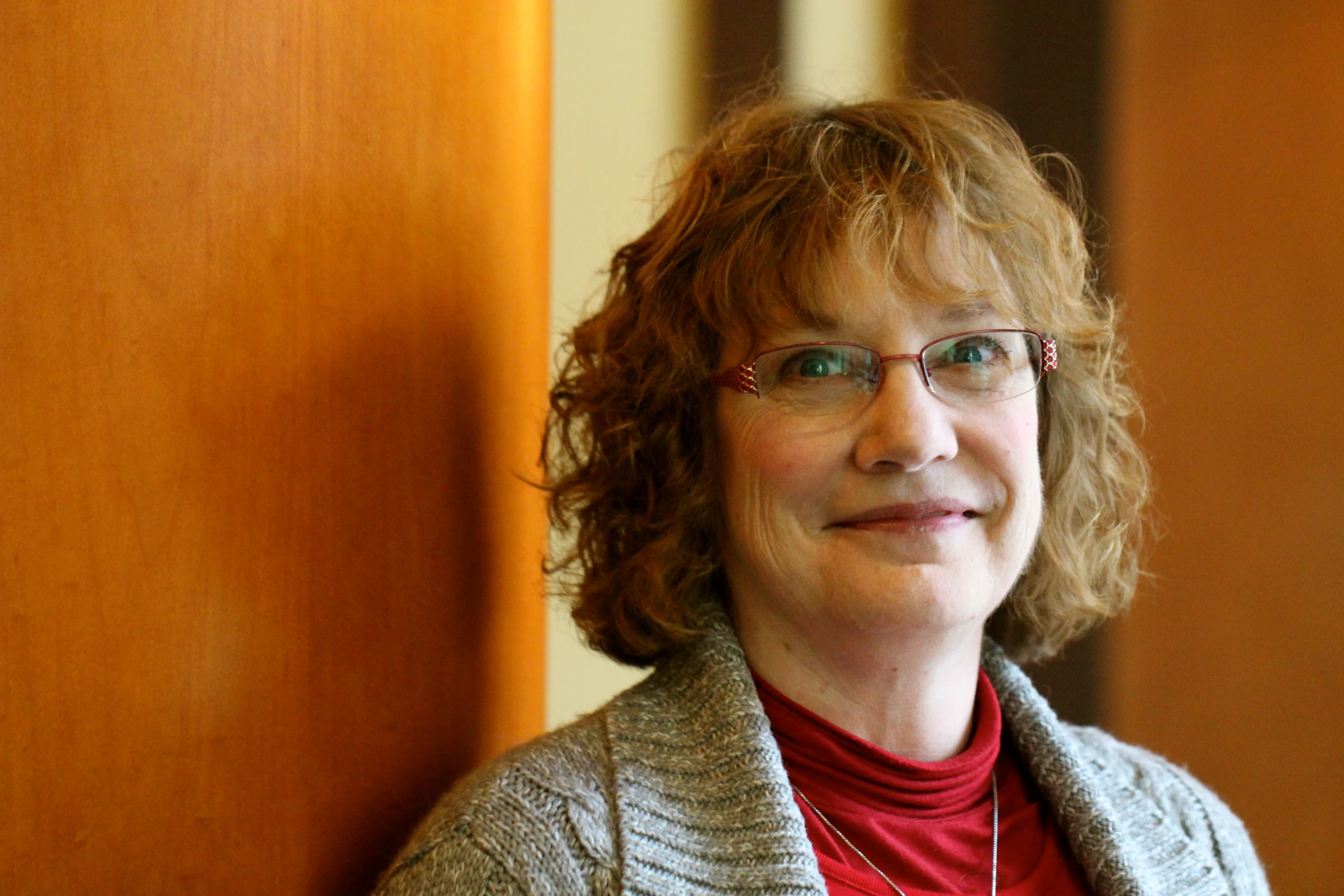 a woman in glasses smiles as she stands near a wooden wall