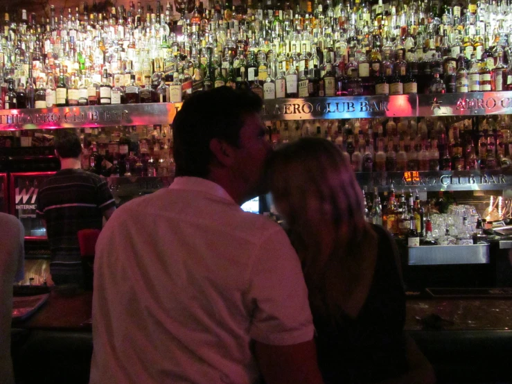 man and woman are at a bar with various liquor bottles on the wall behind them