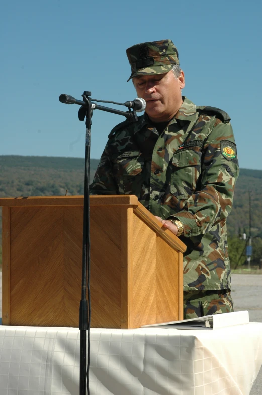 a military officer in camouflage stands at a podium with a microphone