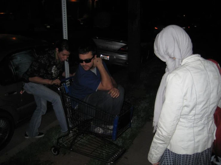 two men standing around in front of a car with a man sitting on the basket