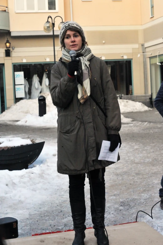 a woman standing on a sidewalk, dressed in winter attire, is holding her cell phone and looking away from the camera