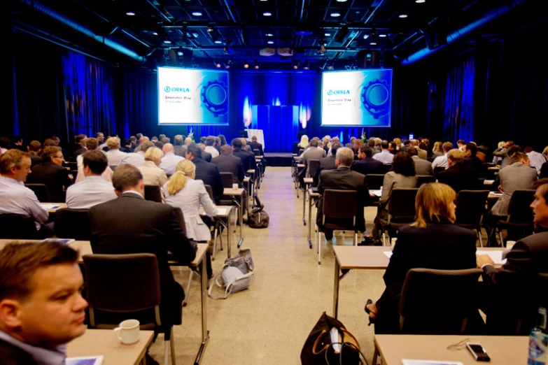 an audience looks at speakers and screen with screens in the back