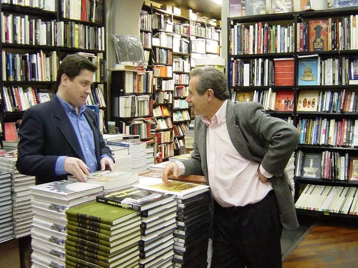 two men are talking in a liry with stacks of books