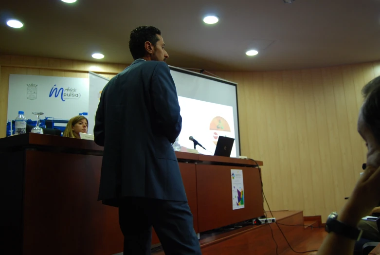 a man giving a speech during a meeting in front of a big screen