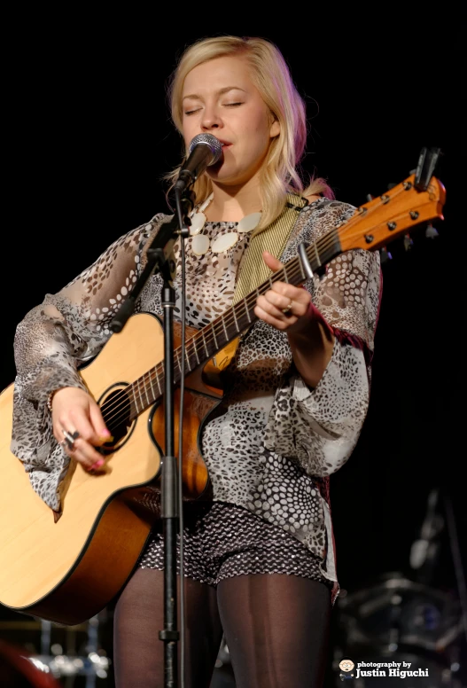 a blonde haired woman wearing tights, with her hands behind her chest, playing a guitar