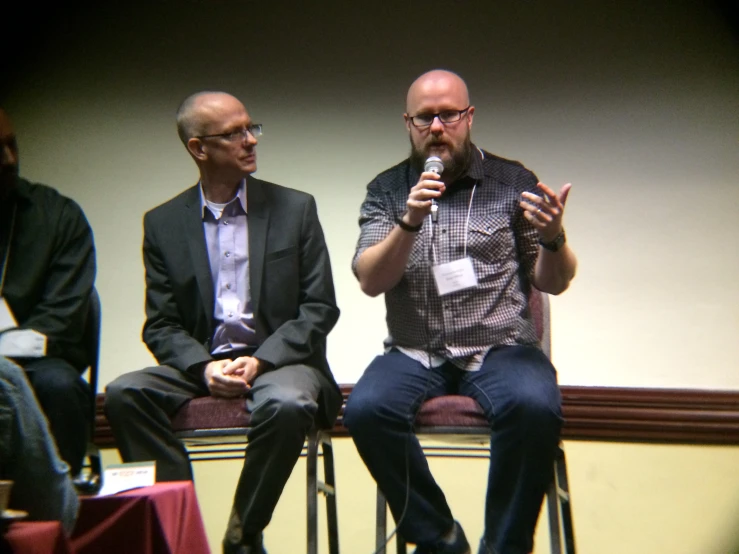 two men sitting on a stage with a microphone and two are holding up their hands