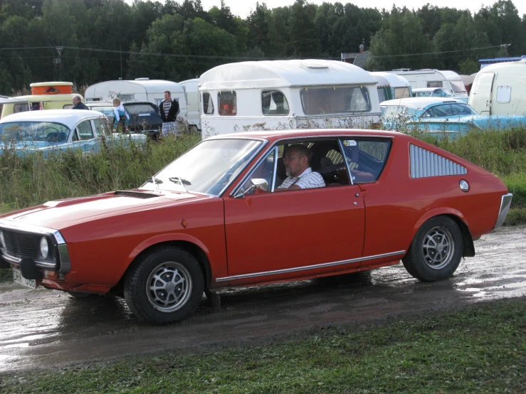 man in small red car stopped in grassy area with campers and trailers in background