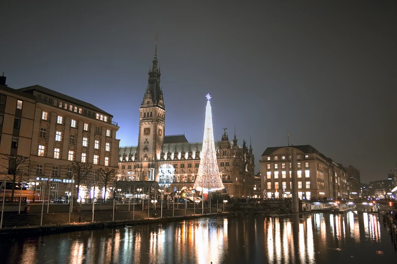 a lighted building next to water with many buildings behind it