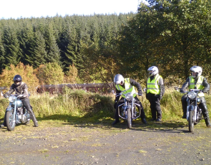 three motorcycle riders, one in safety gear, standing near some trees