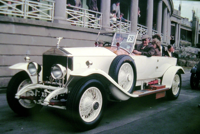 a vintage car is parked in front of a building with people standing on a balcony and two men sitting