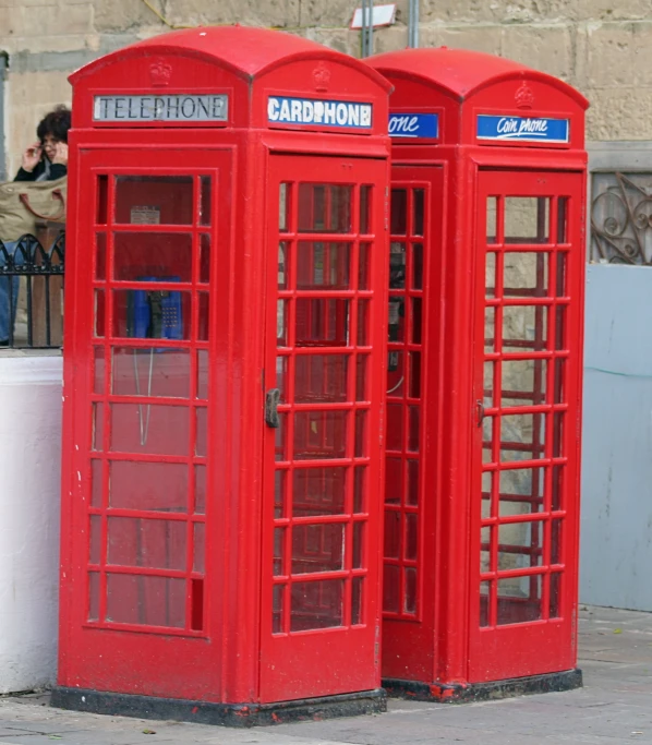 two red phone booths on a sidewalk with people sitting