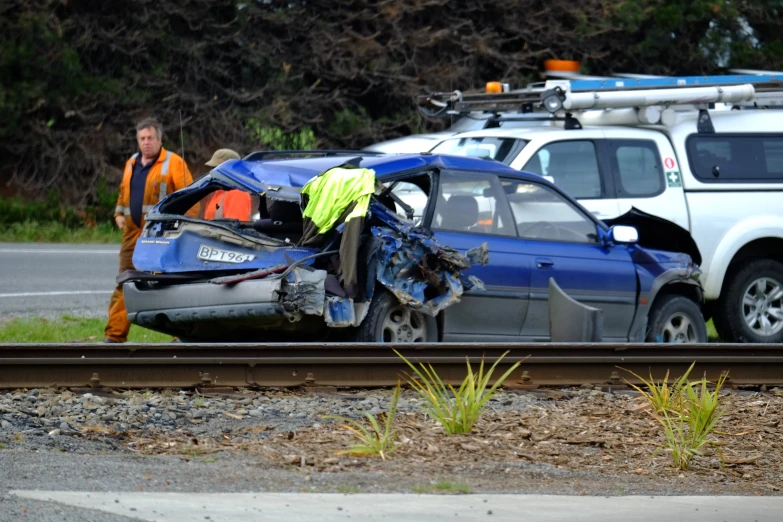 a crashed car sitting on the side of a train tracks next to people walking