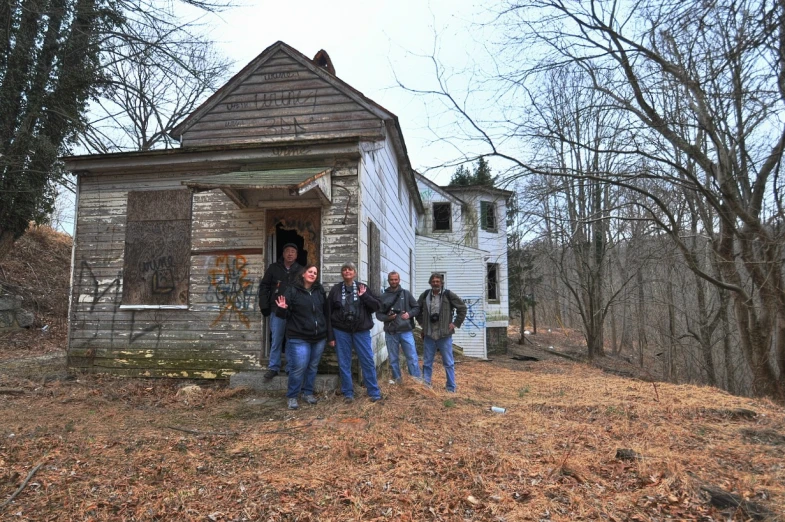 five men stand in front of an old house in the woods