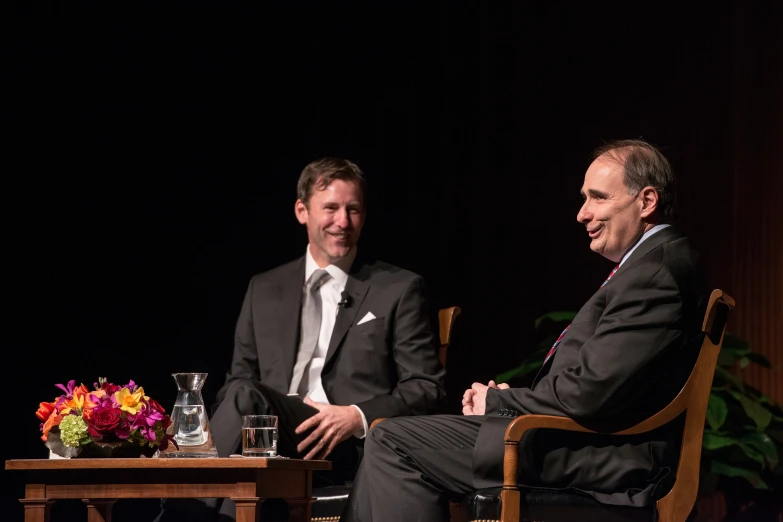 two men in suits sitting down talking and smiling