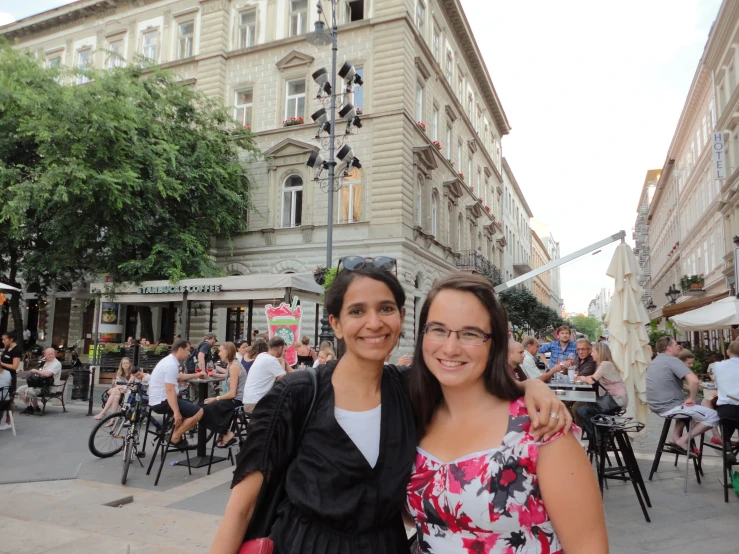 two women standing side by side in front of a building with many people seated at tables in it