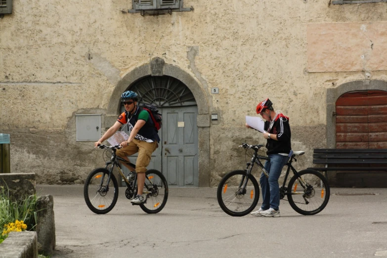 two men on bicycles looking at paper while talking on the street