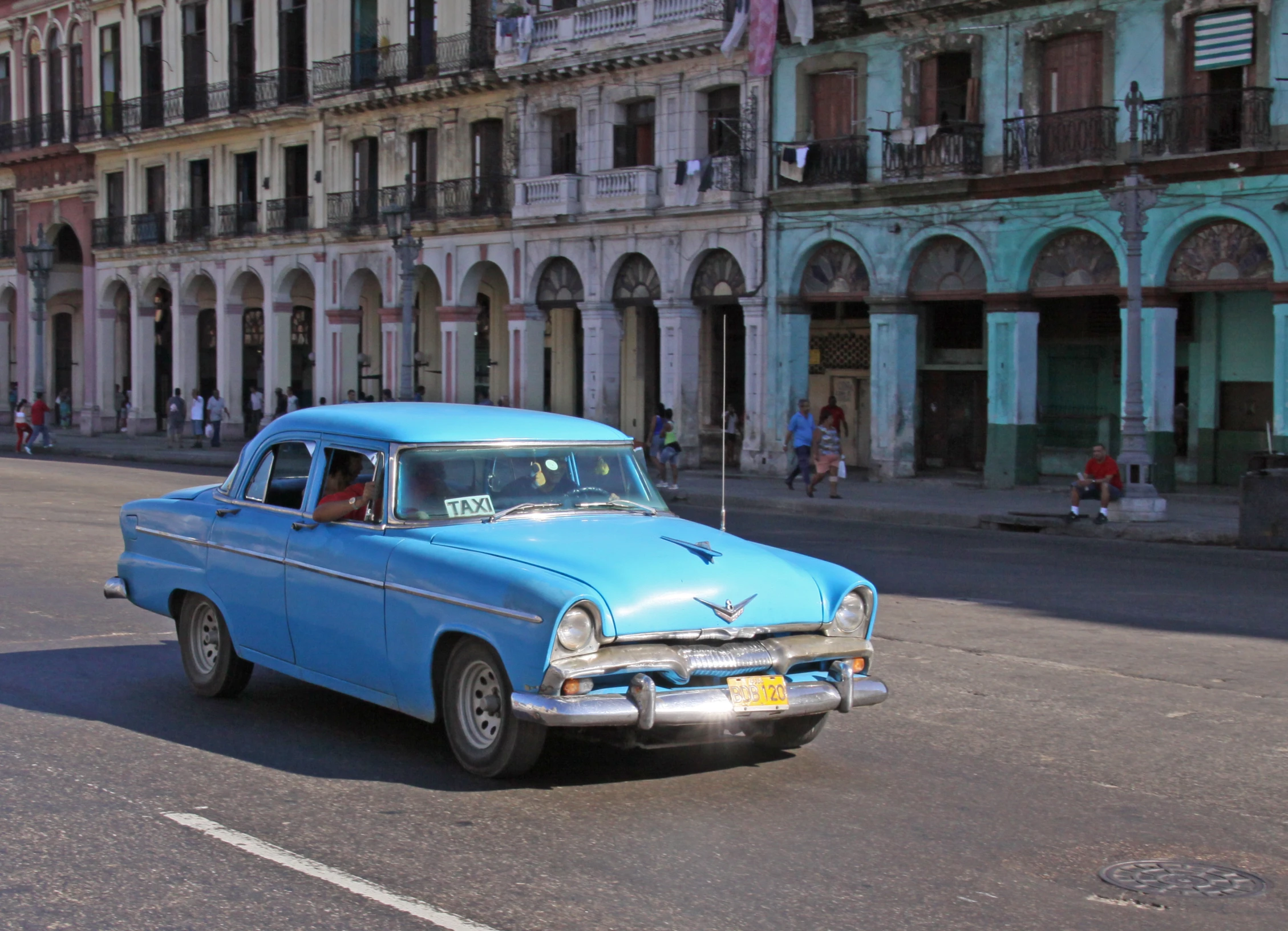 an old blue car sits in front of the buildings on a road