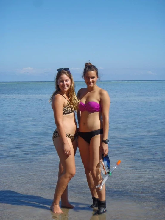 two young ladies in bathing suits stand on a beach by the water
