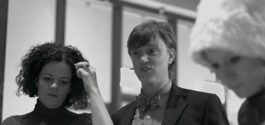 two people wearing formal wear talking while one uses a hair dryer on their head