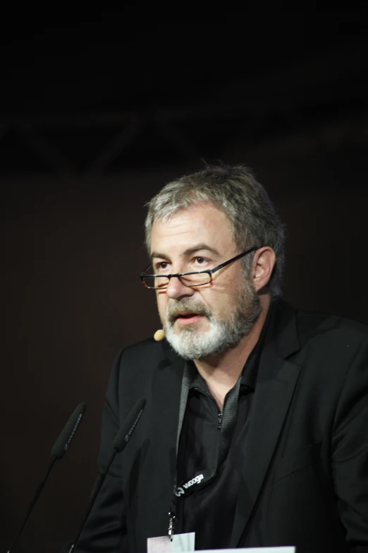 a man with glasses and a black jacket sitting in front of a microphone