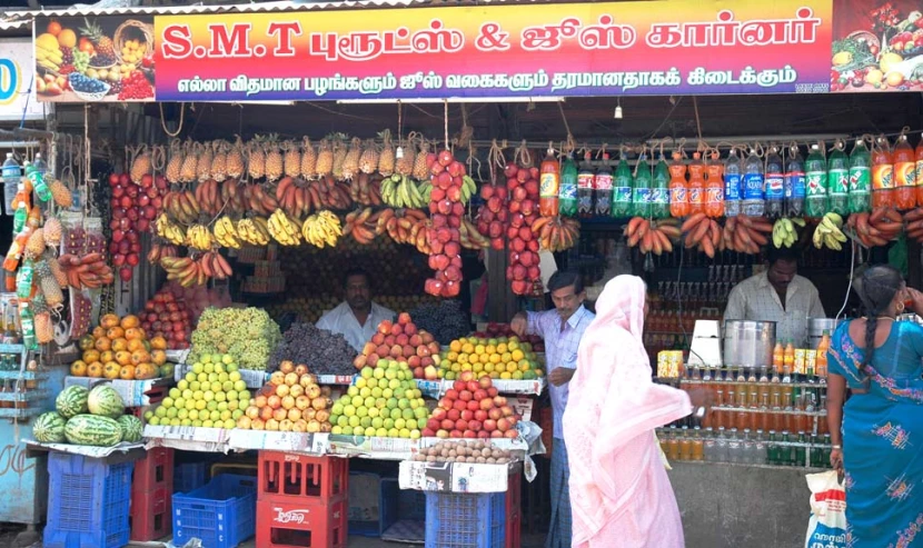 people standing in front of fruit stand with bananas, melons, oranges and other produce