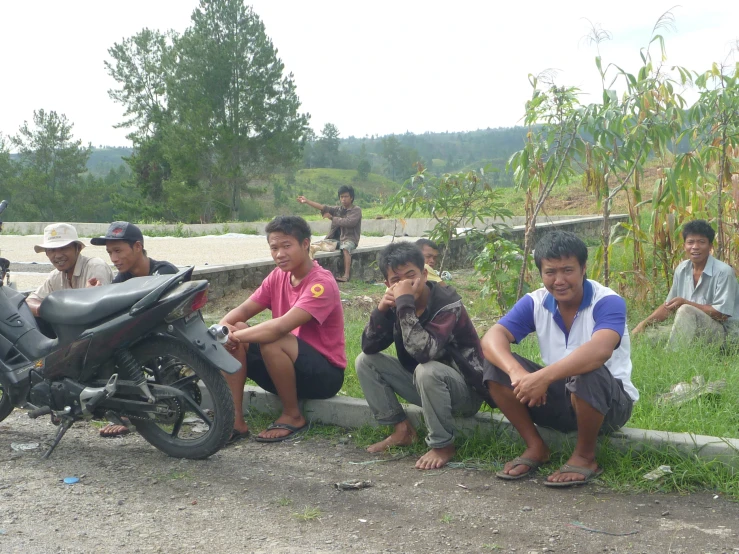 four men with motorbikes and one bike sitting beside a fence