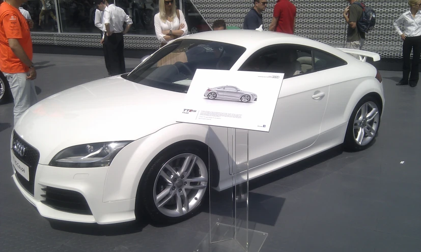 white car on display in a black background with people walking near by