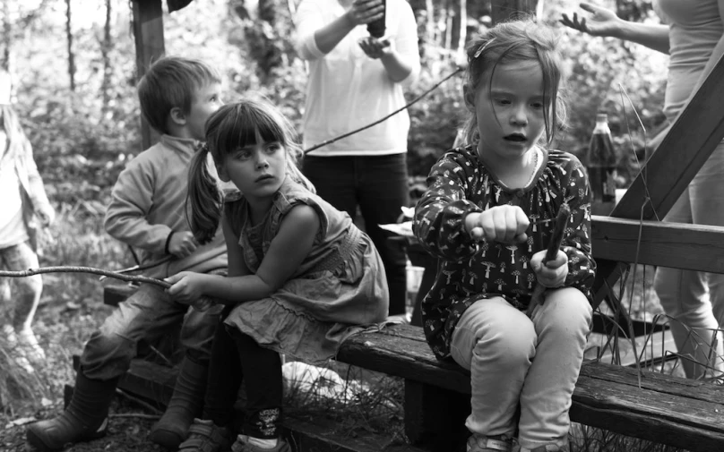 two young children are sitting on a bench with other adults and some children playing