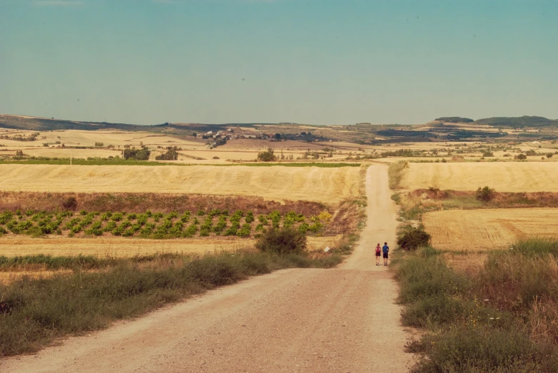 two people are walking up the dirt road towards a field