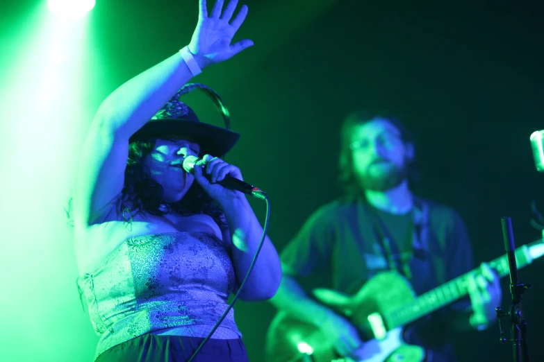 a woman is singing into the microphone while standing with her hands up as she holds an arm above the soundboard and an electric guitar in front of a man