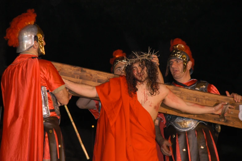 three men dressed in roman garb carry a wooden board