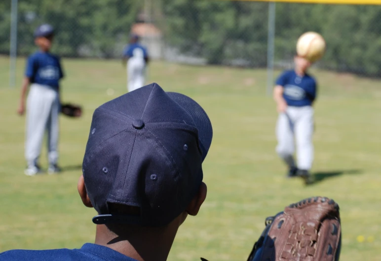 the baseball player wearing a ball cap watches as the pitcher and catcher prepare to catch it