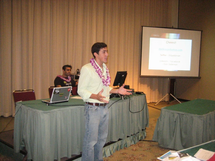 man standing next to an open laptop on a table with two others in front of a projection screen