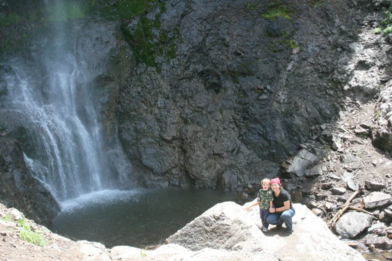 a woman in a hat sits on the edge of a cliff, near a waterfall