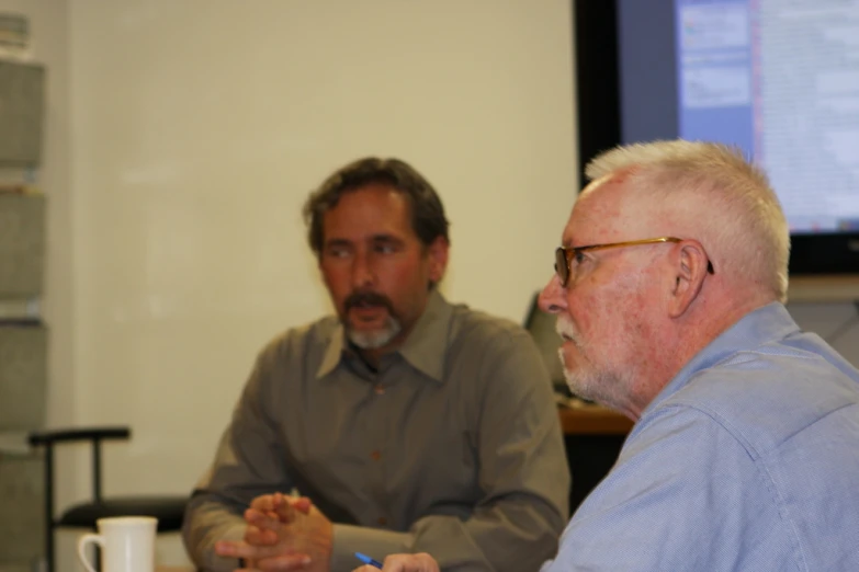 two men sitting at a table in front of a projector screen