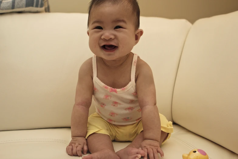 a small baby wearing a white tank top sits on a couch