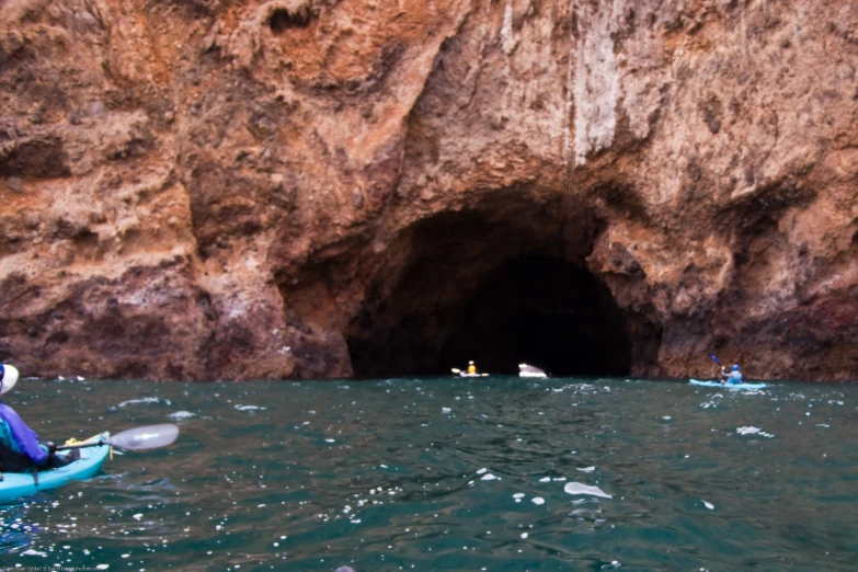 three kayakers paddling past a large cave that has ice chunks on it