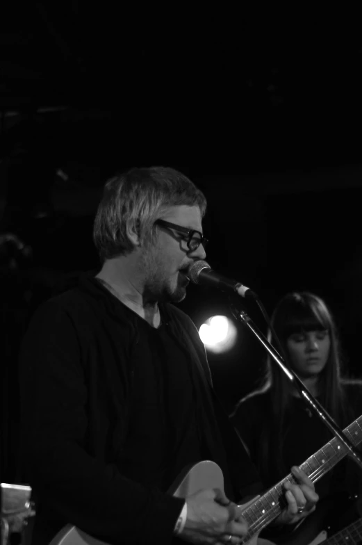 man with glasses singing into microphone and guitar on stage