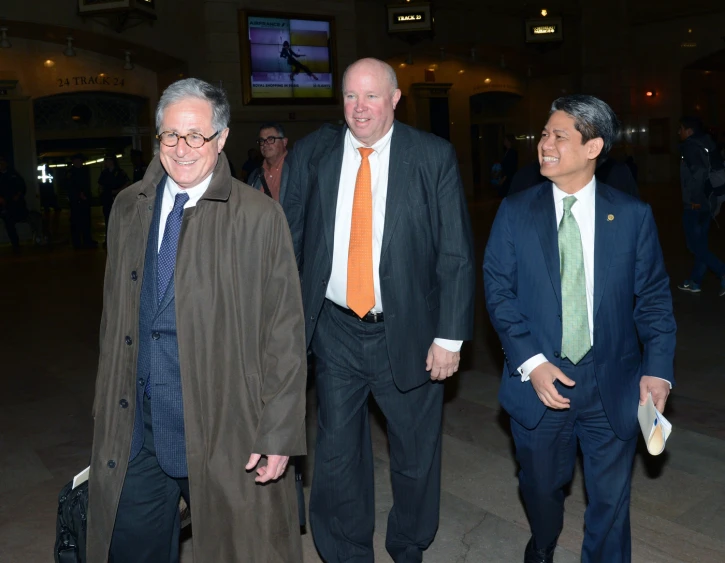 three men wearing suit, two in suits and one in orange tie walk through a hallway