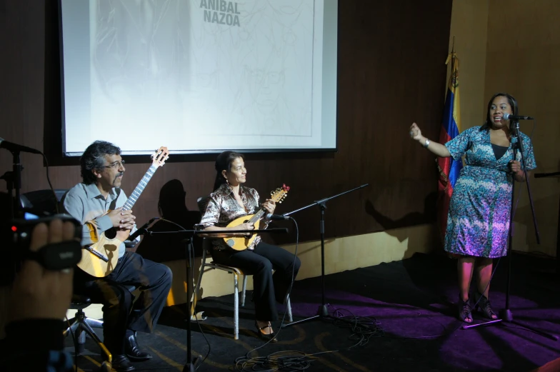 two men playing guitars and singing into microphones as a woman sits near a mic