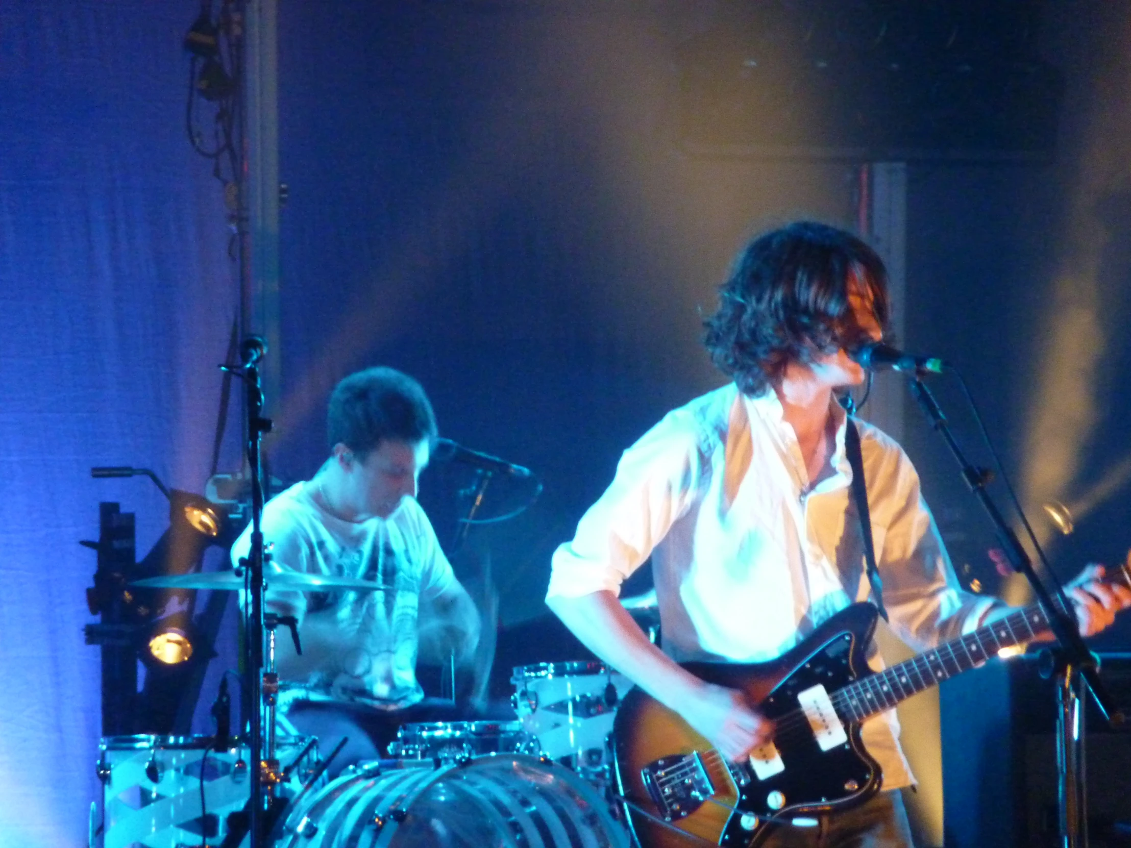 two young men playing guitar while another man in white shirt watches them