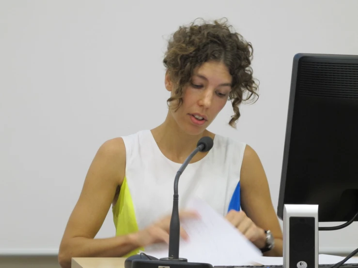 the woman in white shirt is holding papers while the man looks at the screen